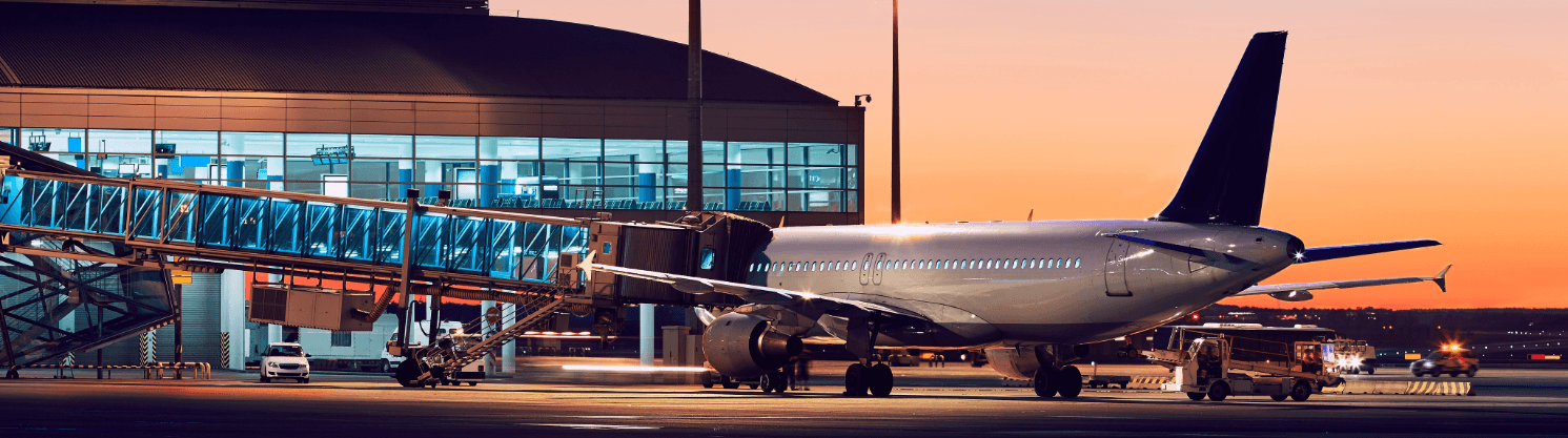 Flugzeug an einem beleuchteten Terminal bei Sonnenuntergang, Fluggastbrücke angedockt, Vorfeld am Abendhimmel.