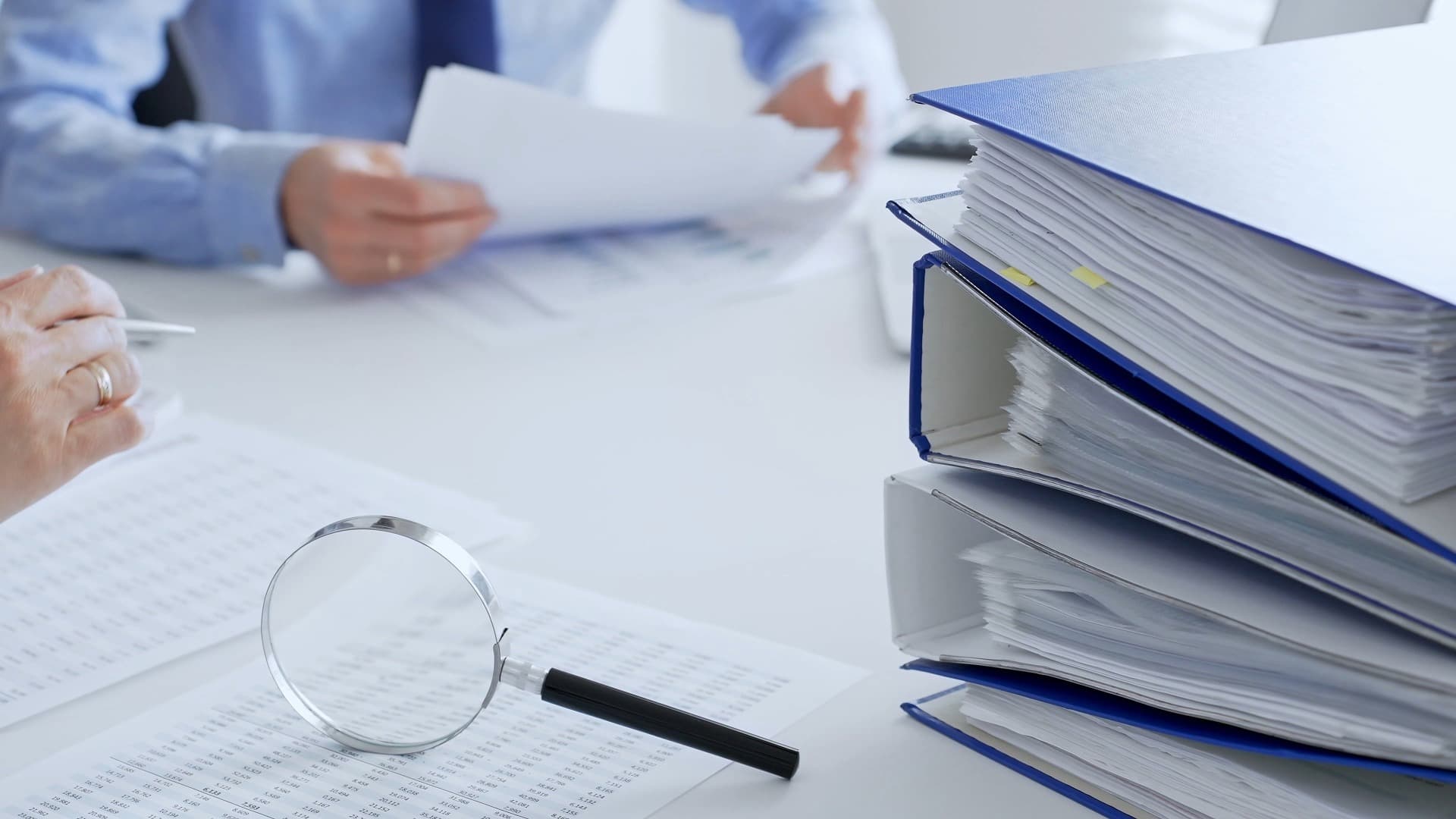Person reviewing prior damage reports with magnifying glass next to stack of blue binders in office