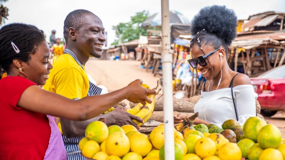 Three Nigerians are standing at a market stall selling bananas and lemons
