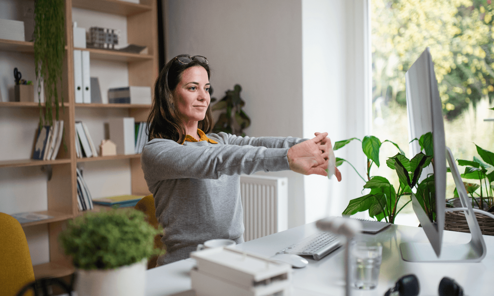 Woman stretching wrists in bright home office after moving with desk, computer, and plants. Setup for healthy working from home.