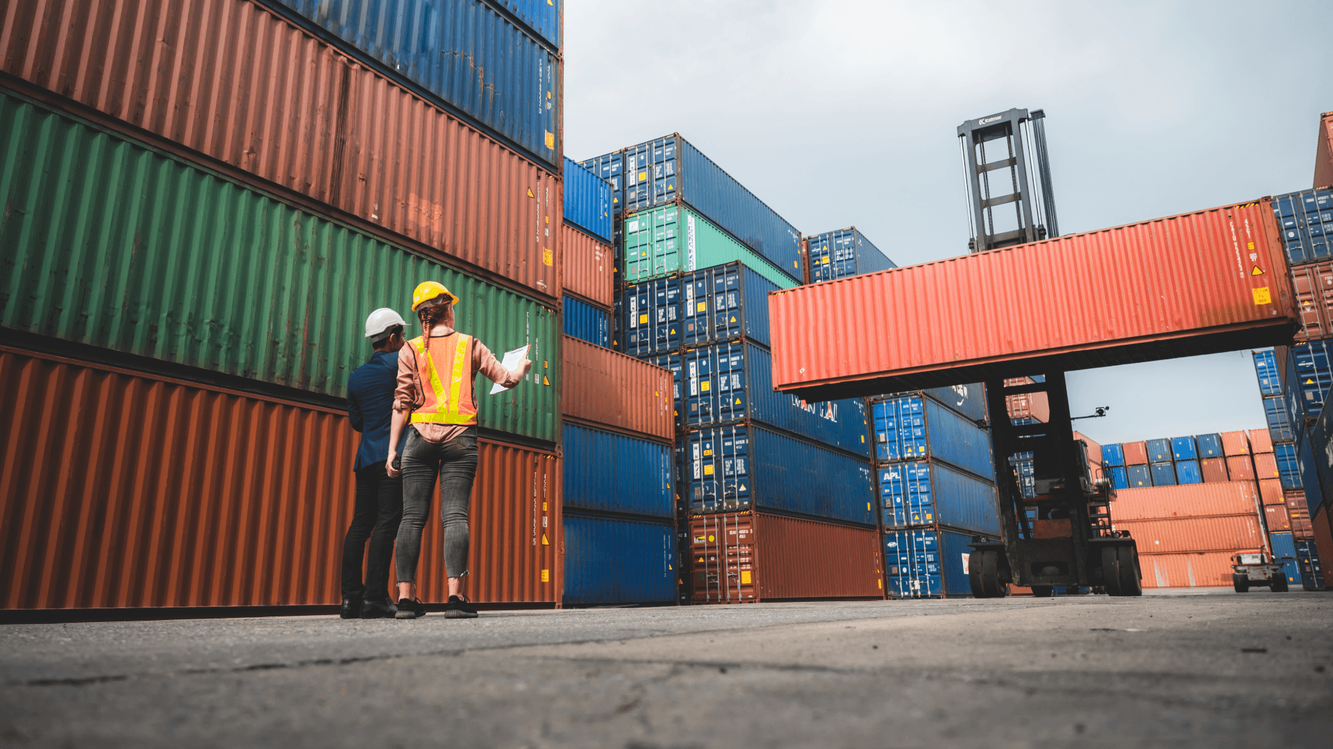 Two port workers stand in front of stacked colorful containers. A forklift moves an FCL container.