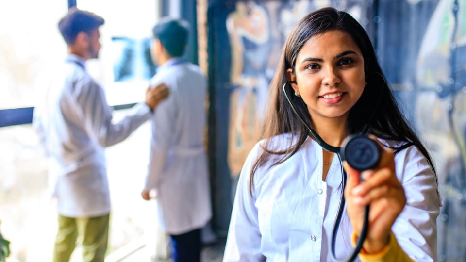 A Moroccan doctor holds a stethoscope up to the camera; in the background, two other doctors are talking