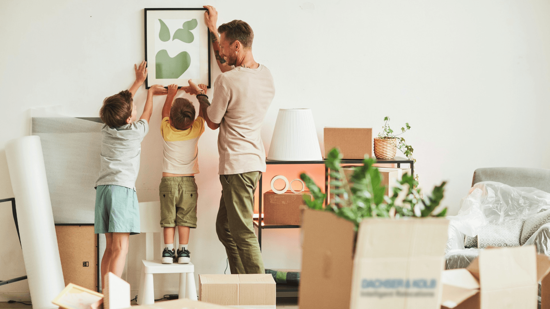 Familie hängt Bild in neuer Wohnung auf, Umzugskartons im Raum – Günstig umziehen