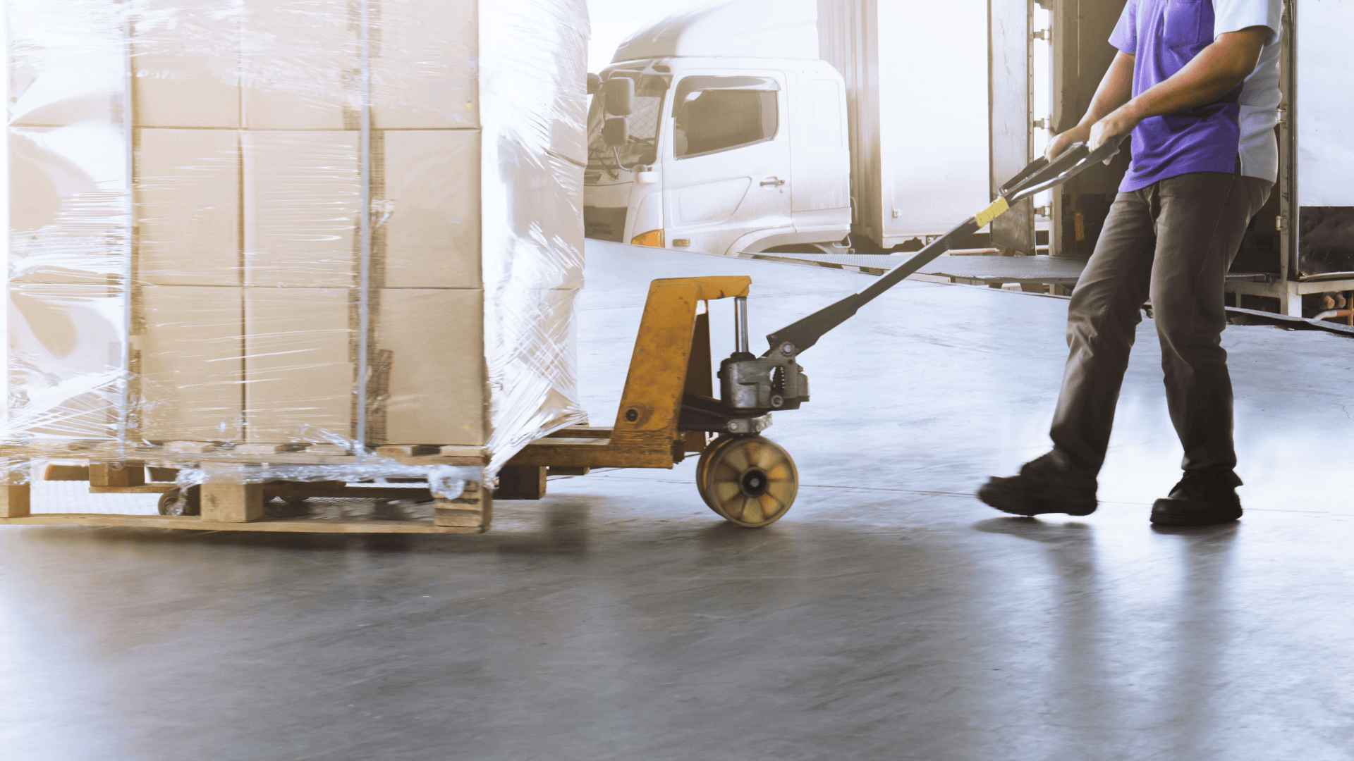 Worker pulling a pallet jack with wrapped cardboard boxes. No wooden crates visible. Warehouse and logistics.