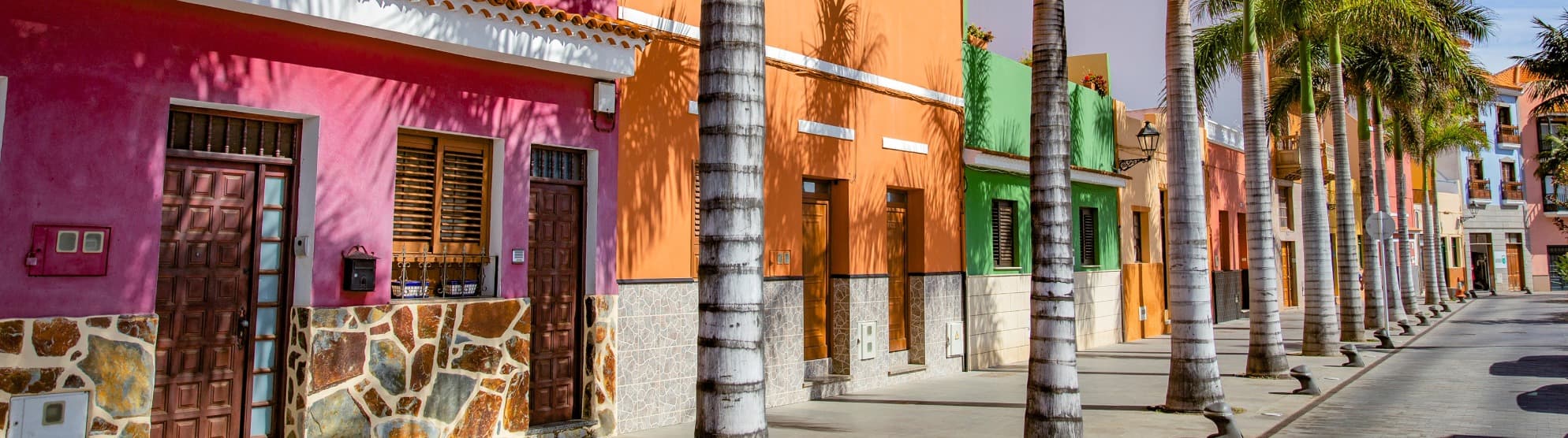 Bunte Häuserfassaden in Pink und Orange an einer mit Palmen gesäumten Straße auf Teneriffa, genutzt als Symbolbild für den Versand von Umzugskartons mit DACHSER & KOLB.