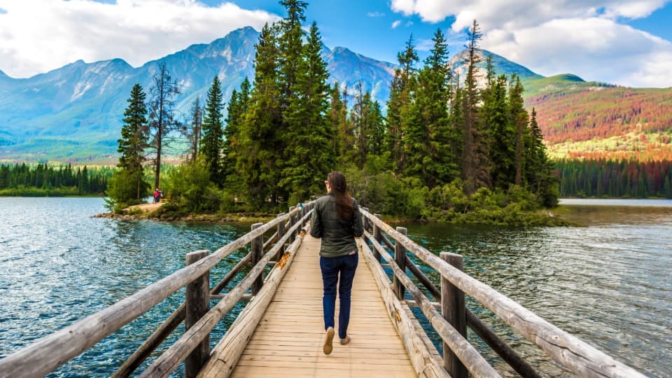 A Canadian woman walks across a wooden bridge over a body of water toward an island with a small forest.