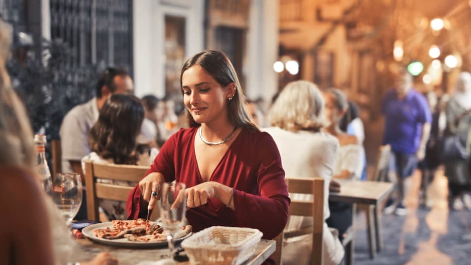 A smiling woman is eating pizza in the evening at a bustling outdoor restaurant – a scene that epitomizes the Mediterranean lifestyle and Maltese culture.
