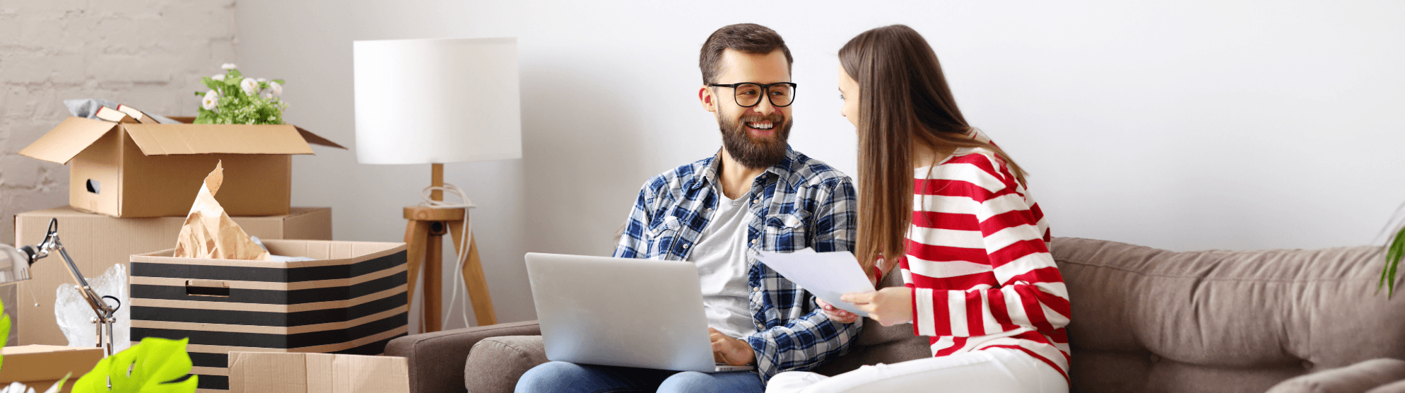 Happy couple planning move with laptop and documents for transit insurance. Moving boxes in background.