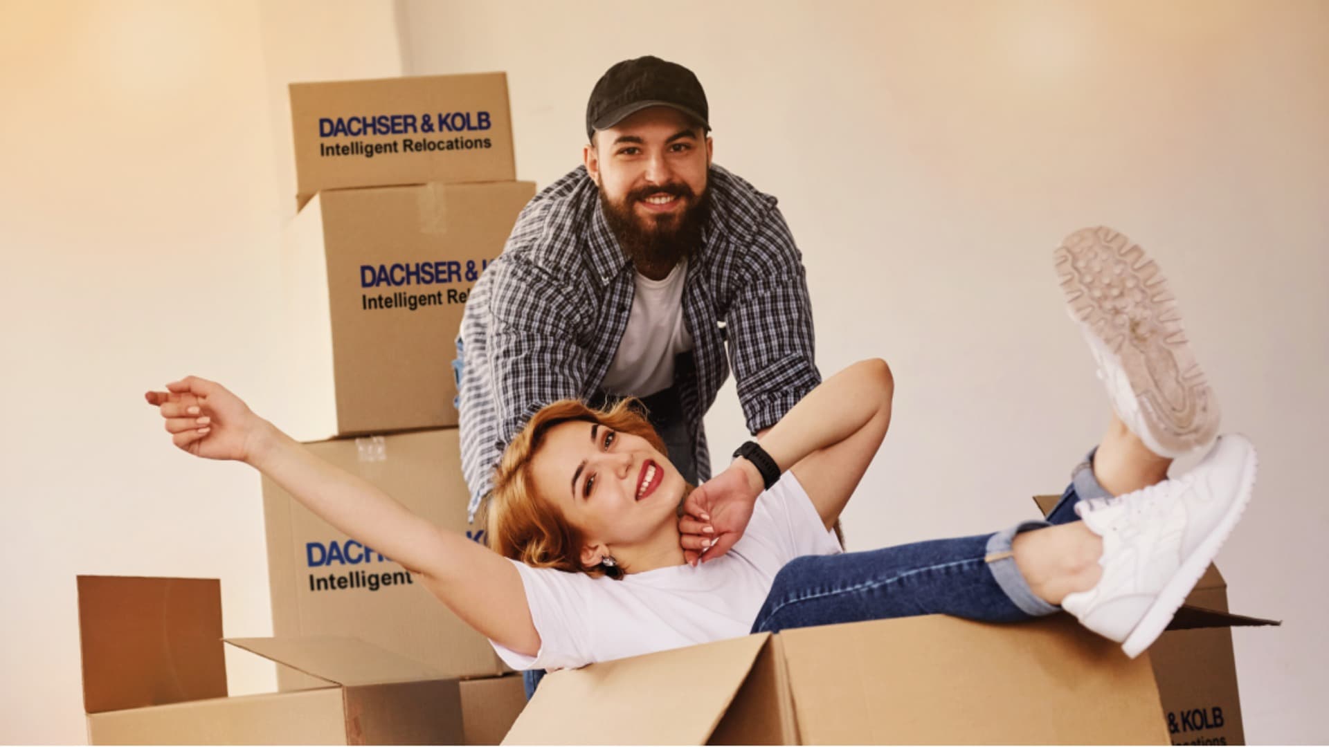 Smiling couple moving, woman sitting in DACHSER & KOLB moving box – Quality and reliability with FIDI certification.