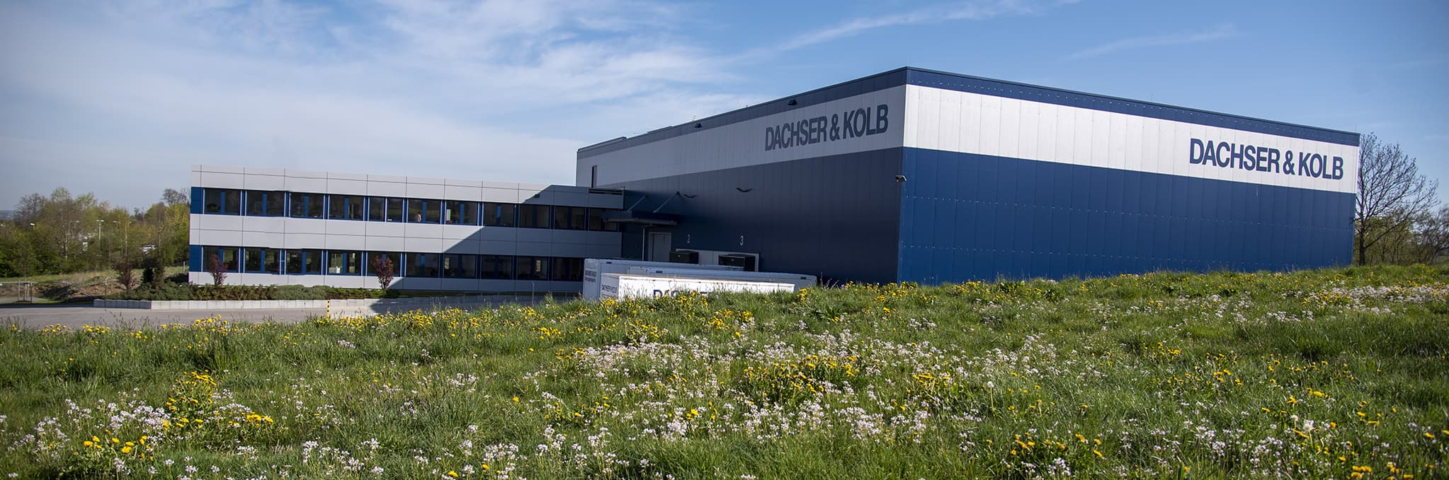 Modern building of the moving company DACHSER & KOLB with a warehouse and office section, surrounded by green meadow.