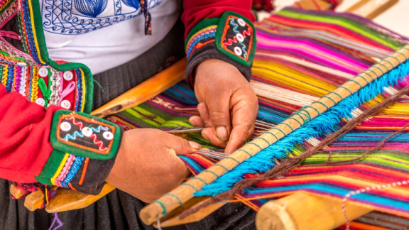 A Peruvian man in traditional clothing weaves a colorful cloak