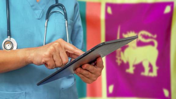 A doctor from Sri Lanka stands in front of the national flag and types something into a tablet