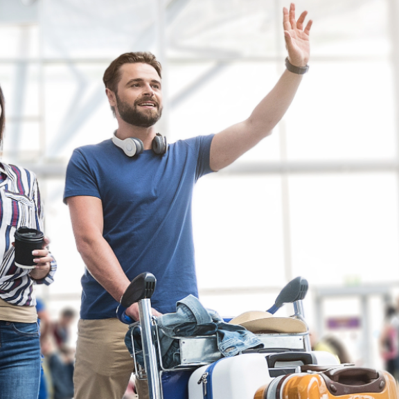 Junges Paar mit Gepäck am Flughafen, winkt fröhlich – bereit für ihren internationalen Umzug. Helle, geschäftige Terminalszene.