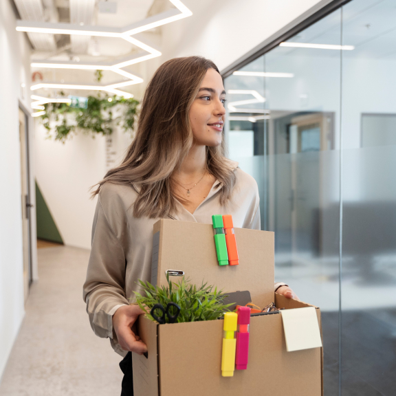 Young woman carrying moving box through modern office – company relocation with personal items.