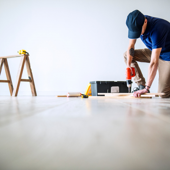 Handyman Service: Man kneeling on wood floor, drilling hole, toolbox and ladder in background.