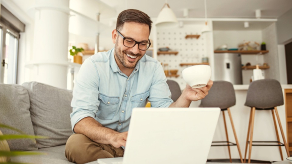 Ein junger Mann sitzt lachend mit einer Kaffeetasse auf dem Sofa und nutzt den Online-Rechner von DACHSER & KOLB für sein Umzugsangebot.