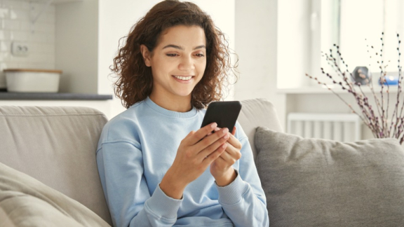 Eine junge Frau sitzt entspannt auf dem Sofa und nutzt ihr Smartphone für die digitale Volumenermittlung oder Online-Besichtigung ihres Umzugs mit DACHSER & KOLB.