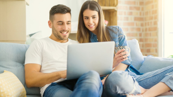 Ein glückliches Paar sitzt entspannt auf dem Sofa vor Umzugskartons und nutzt am Laptop die Ratenzahlung für den Umzug mit DACHSER & KOLB.