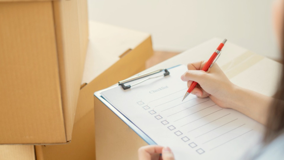 A person ticking off a structured checklist on a clipboard next to moving boxes, preparing for an international relocation with DACHSER & KOLB.