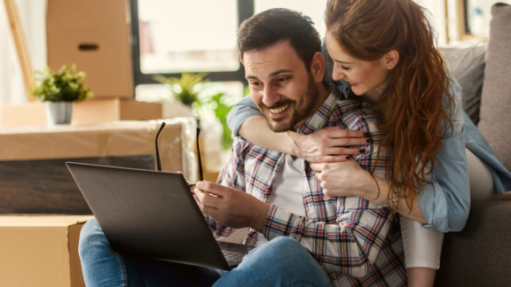 A happy couple sitting with a laptop among moving boxes, using the digital cost calculator for their move to Canada with DACHSER & KOLB.