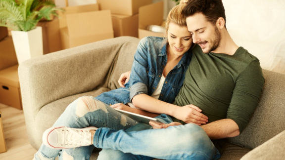 A happy couple sitting relaxed on a couch among moving boxes, calculating their move to the USA using the online calculator by DACHSER & KOLB on a tablet.