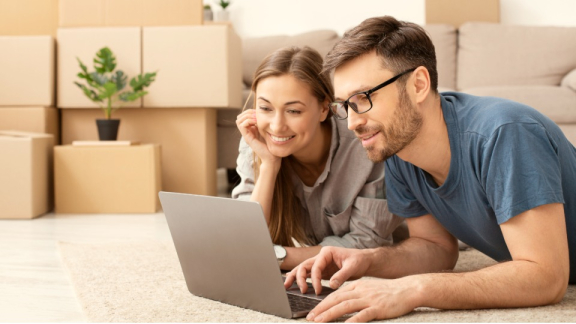 A smiling couple uses a laptop to calculate Brazil moving costs with the digital moving calculator from DACHSER & KOLB, with packed moving boxes in the background.