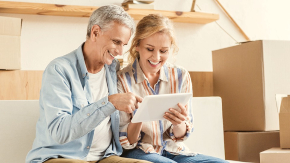 An older couple happily uses a tablet to calculate Thailand moving costs with the digital moving calculator from DACHSER & KOLB, surrounded by moving boxes.