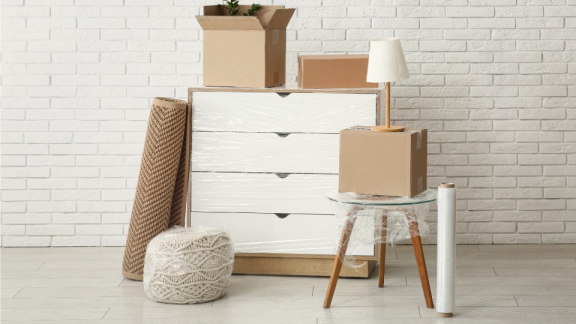 A wrapped chest of drawers, a rug, and packed moving boxes in front of a white wall as typical examples for small volume transport with DACHSER & KOLB.