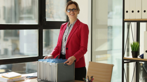 A smiling businesswoman in a red blazer organizing files in an archive box in an office, representing commercial small volume transport with DACHSER & KOLB.