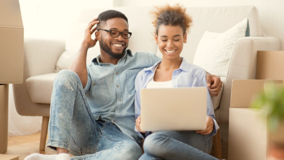 Happy couple sitting between moving boxes with a laptop calculating moving costs using the online calculator from DACHSER & KOLB.