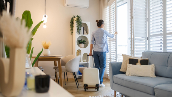 Woman in a new home, opening blinds and pulling a suitcase. Symbolizing relocation service for expats.