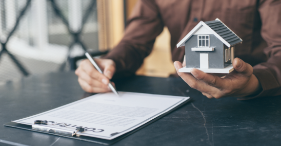 A man signs a contract while holding a house model, symbolizing the Relocation Service for Expats.