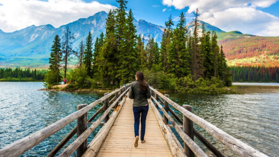 A Canadian woman walks across a wooden bridge over a body of water toward an island with a small forest.
