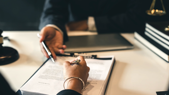 Eine Person unterschreibt einen Vertrag, während eine andere Person einen Stift reicht. Symbolisiert Vertragsabschluss bei Incoterms.