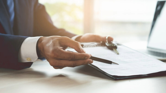 Hand im Anzug zeigt auf Vertragsdokument mit Stift und Autoschlüssel auf dem Schreibtisch